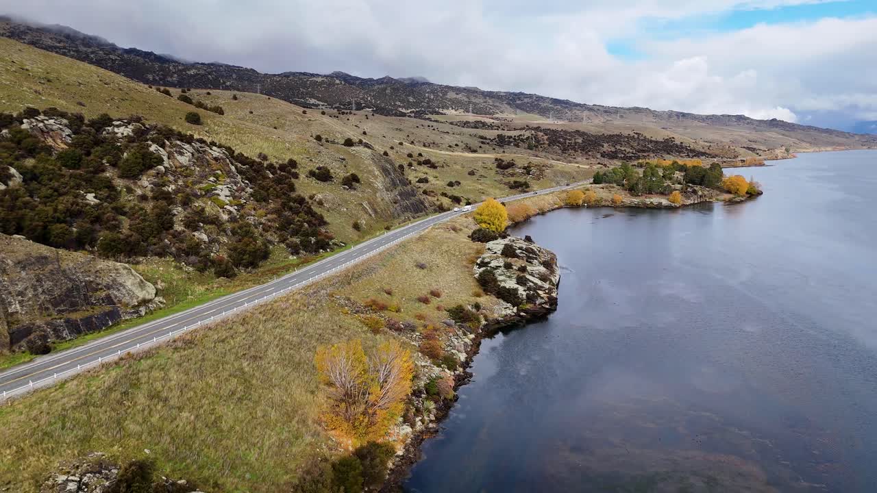 Drone glides above a winding lakeside road bordered by hills and calm water, capturing reflections, natural scenery, and dynamic movement in daylight