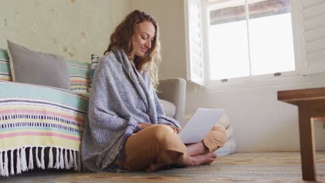 smiling caucasian woman sitting on floor wearing blanket using laptop in sunny cottage living room