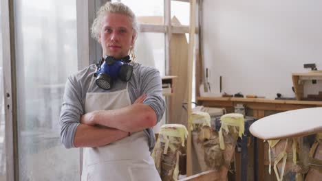 caucasian male surfboard maker wearing a face mask and standing in his studio with his arms crossed