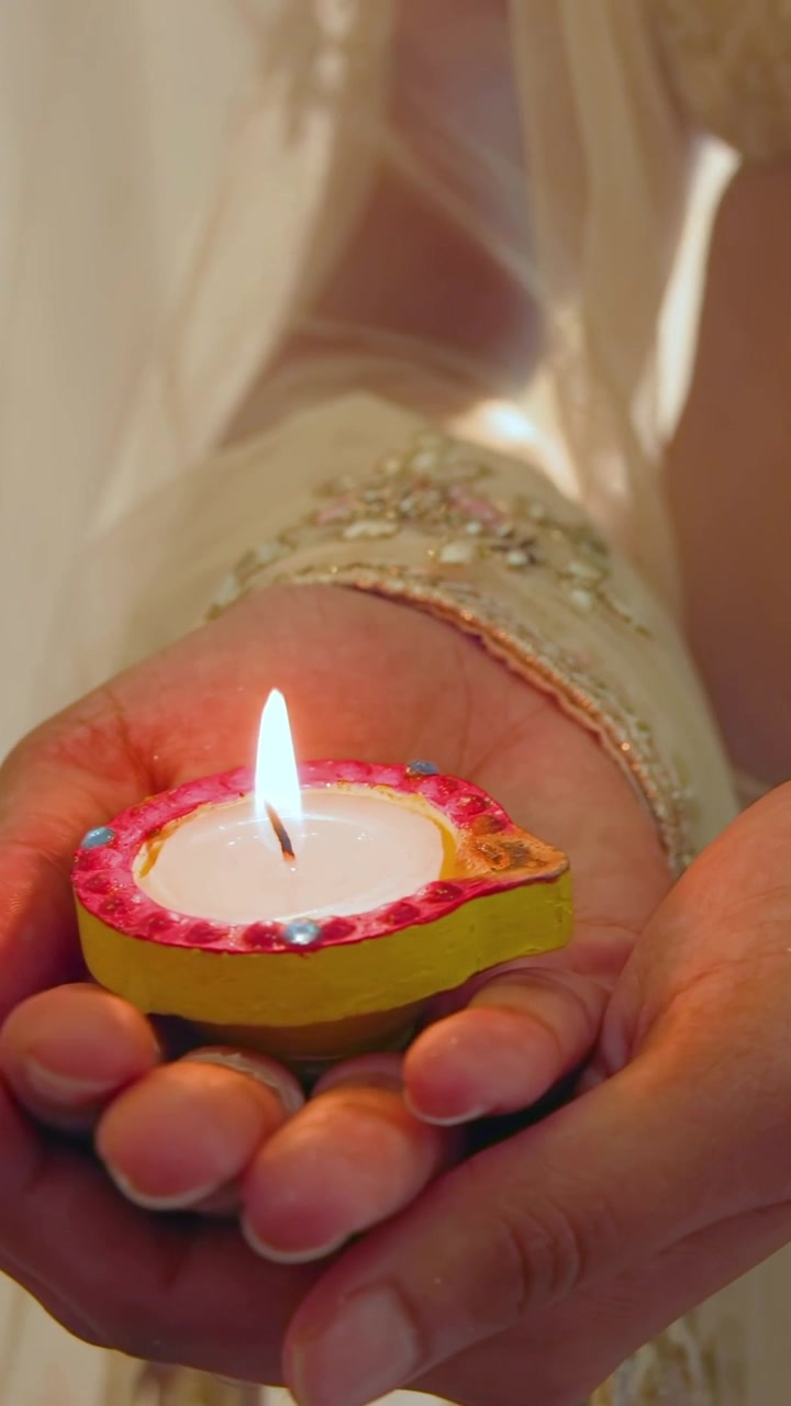 Video verticale: Donna che accende una fiamma a casa, indossando un sari color crema, tenendo una diya con bordo rosa per la puja.