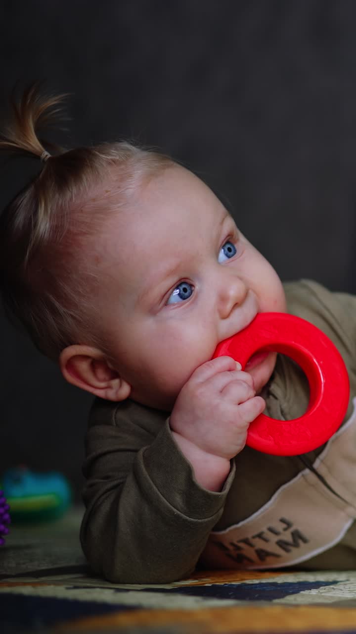 Infant child in a romper lies on the floor. Cute baby picks a toy and takes it to his mouth. Blurred backdrop. Vertical video