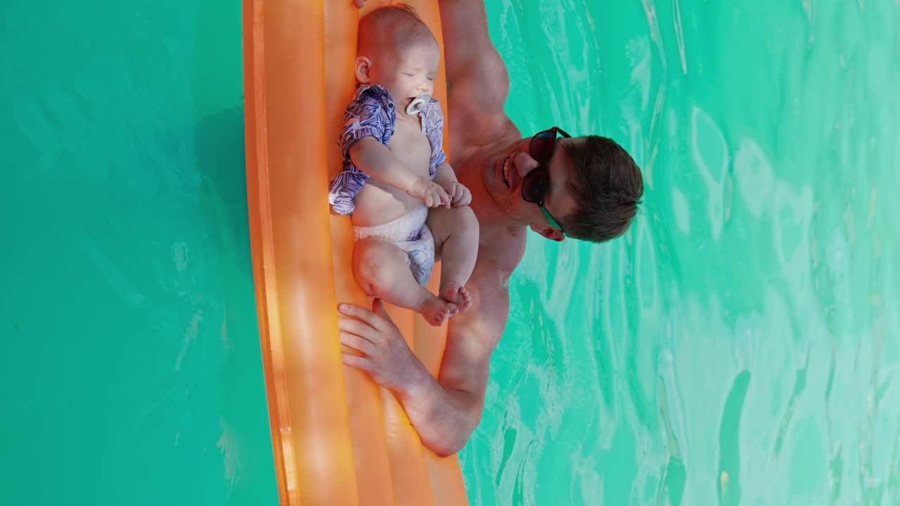 Caucasian baby in diaper and shirt with pacifier in mouth lies on the inflatable mattress. Father is standing at the mattress in the pool and kid cries. Vertical screen.