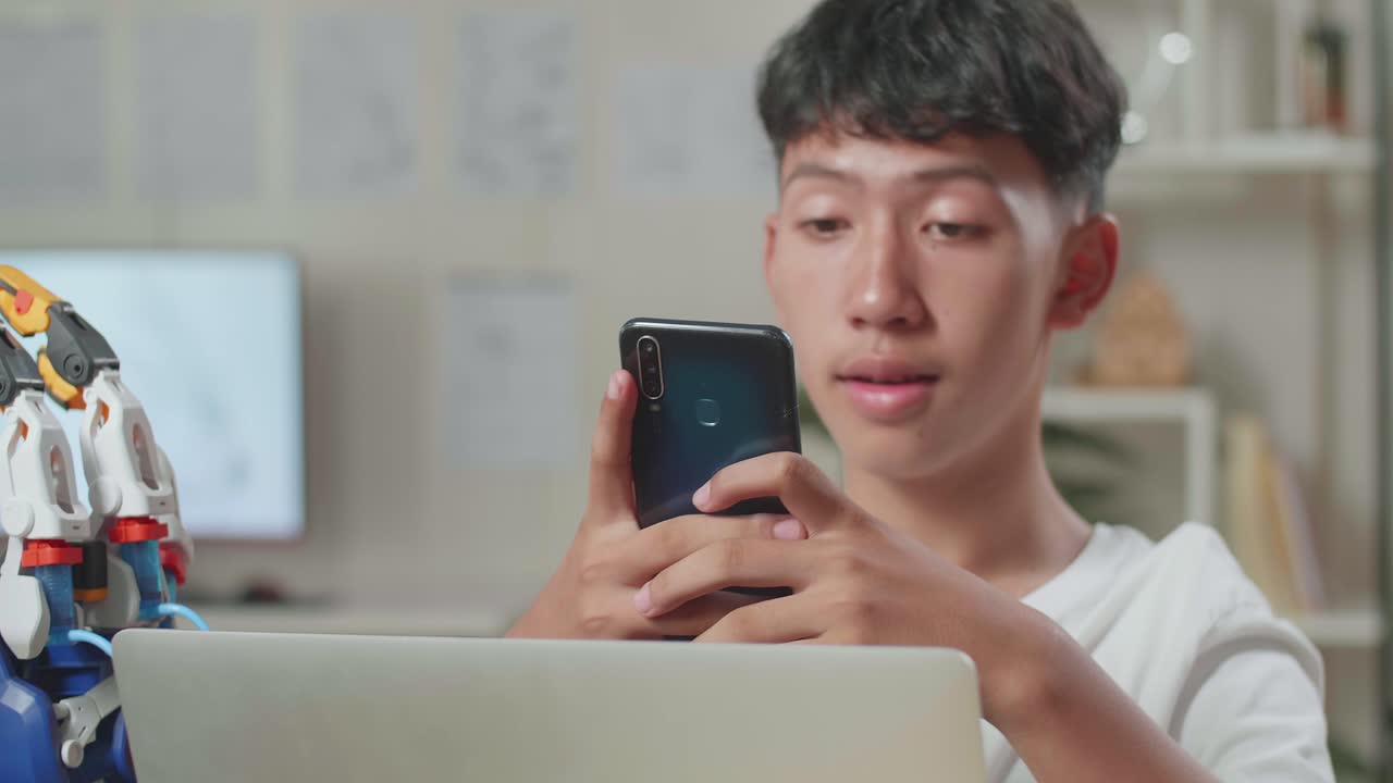집에 있는 노트북에서 사이보그 손으로 작업하는 동안 스마트폰을 사용하는 아시아 소년의 클로즈업