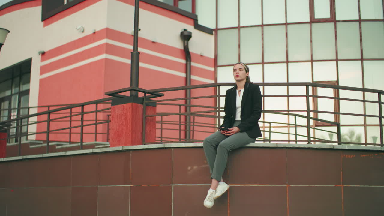 Lady in black blazer and white shirt seated on fence in urban area holding phone, waiting for someone, legs crossed at ankle, modern glass building in background