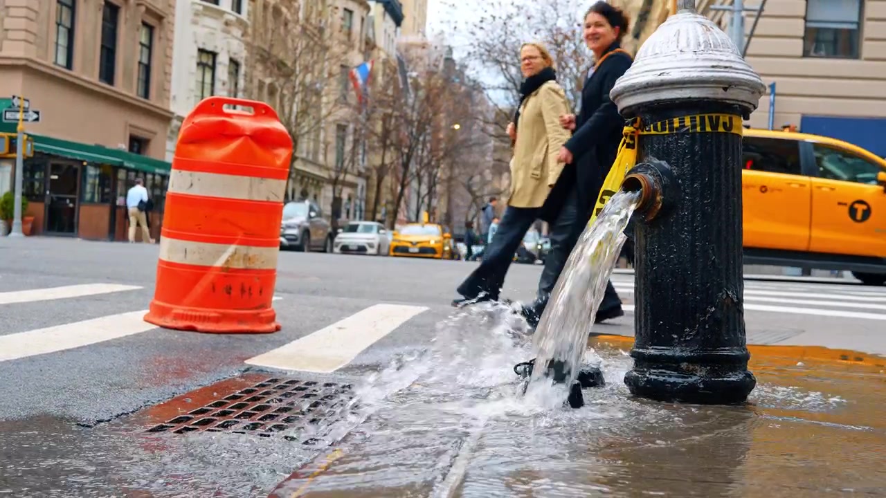 New York, USA, 18 marzo 2026: Acqua che sgorga da un idrante antincendio nero in una strada di New York City. Idrante antincendio aperto che scorre nel canale di scolo stradale vicino a un barile arancione e un taxi giallo a New York City