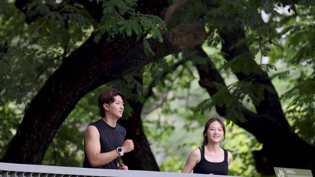 黒いスポーツウェアを着た男女が、緑豊かな公園の木々の下にある橋をジョギングしています。