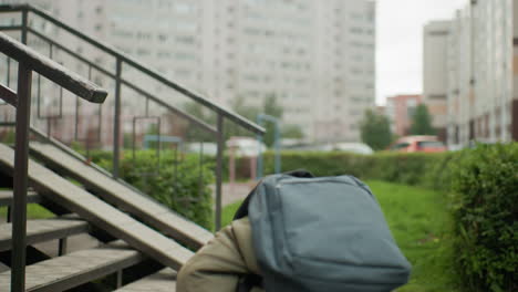 calm kid with backpack carrying book walks through outdoor area toward bench preparing to sit and read with focused expression surrounded by grass, playground frames and urban buildings