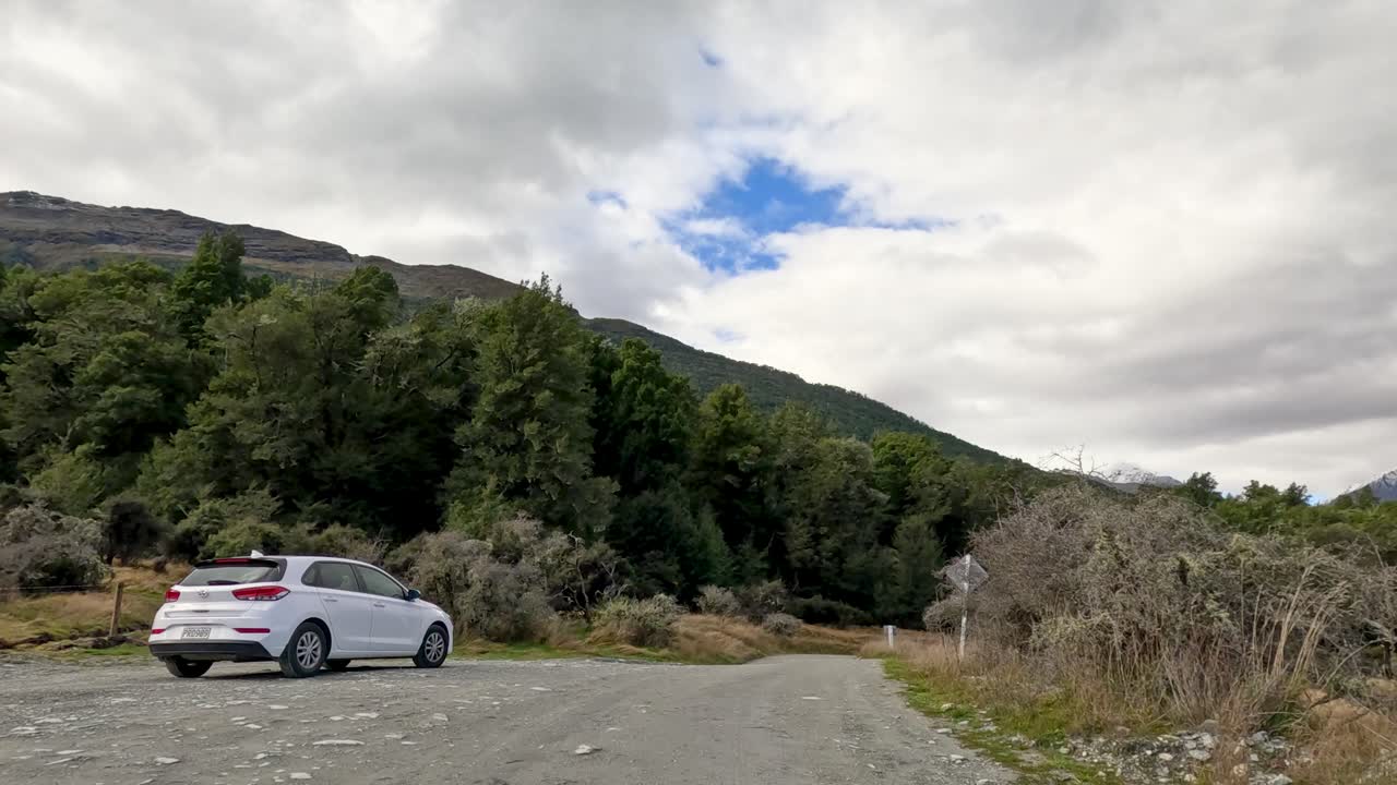 White sedan drives along gravel road toward forested mountains under cloudy sky, wide static shot
