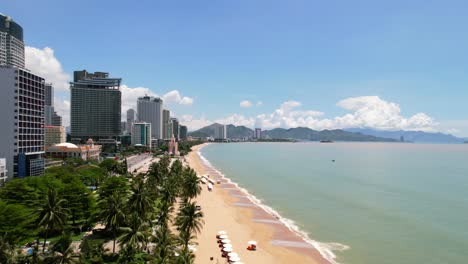 Luftaufnahme-Aus-Geringer-Höhe-Einer-Tropischen-Strandstadt-Mit-Weißem-Sand-In-Asien-Mit-Weißen-Sonnenschirmen-Für-Touristen-Und-Hohen-Hotels-Mit-Blick-Auf-Das-Türkisfarbene-Meer-An-Einem-Sonnigen-Tag-In-Nha-Trang,-Vietnam