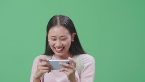 흥분 된 아시아 여성 학생 이 녹색 스크린 스튜디오 에서 는 동안 모바일 게임을 하고 있다