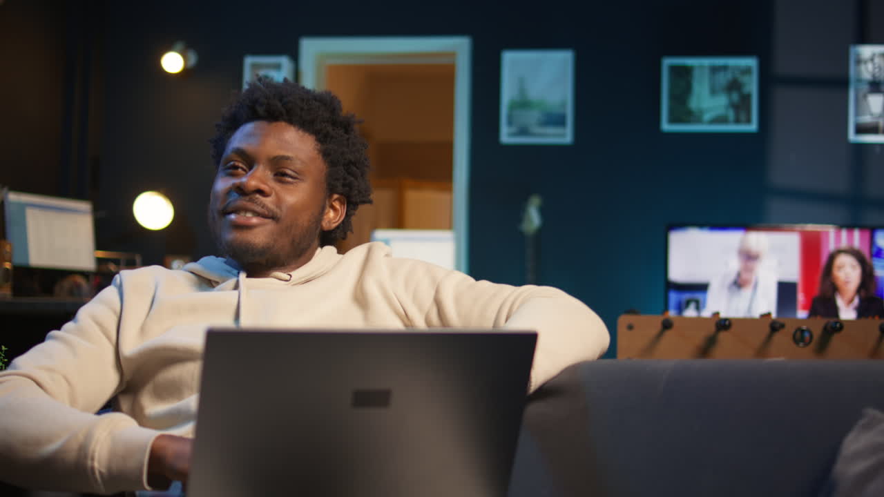 Cheerful African American man teleworking on his laptop in a stylish living room. Remote worker balancing comfort and productivity in a modern flexible workspace. Comfortable loft studio. Camera B.