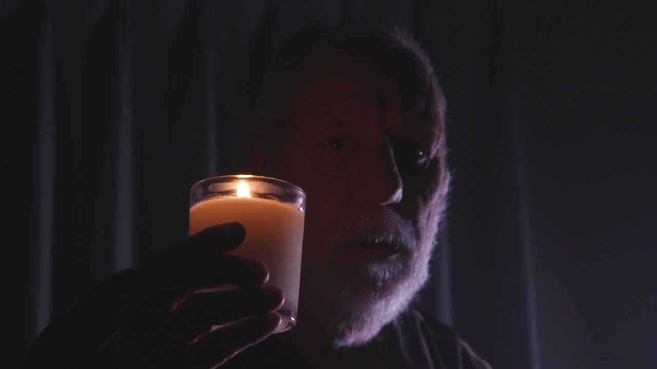 En äldre man håller ett ljus i ett mörkt rum, som kastar ett mjukt sken över hans ansikte. Scenen förmedlar en känsla av ensamhet och eftertanke. Lämplig för teman som reflektion eller introspektion