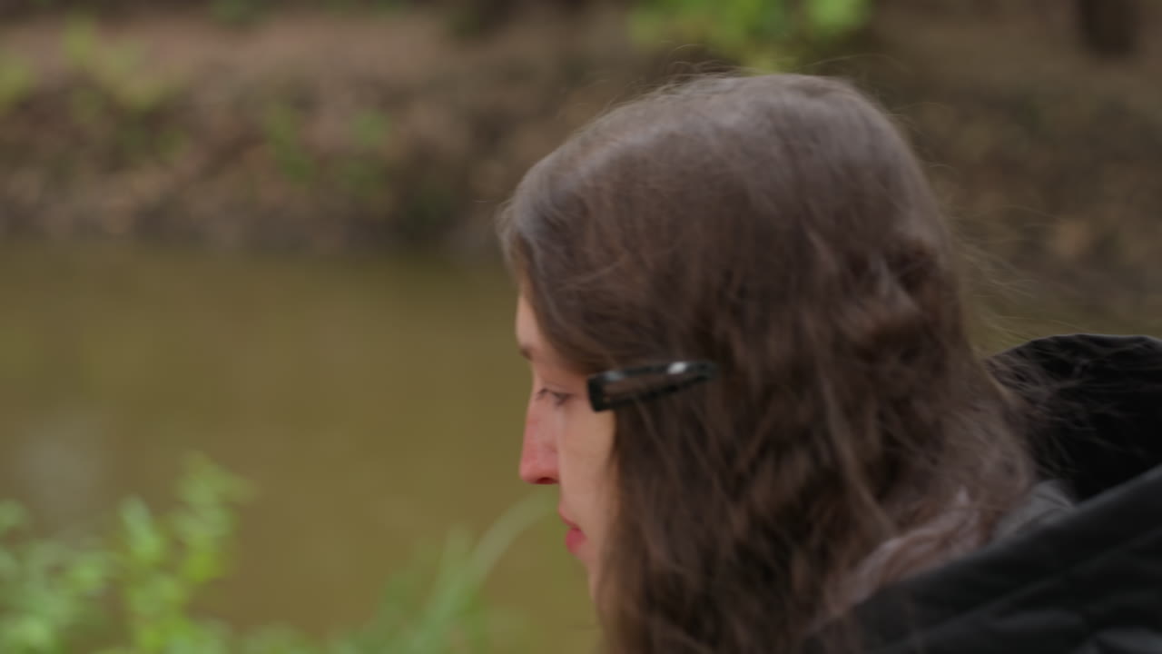 white woman walking riverside pensive profile, student taking reflective stroll along riverbank, hooded jacket with hair clip, muted overcast light, blurred green foliage, calm slow pacing, meditative