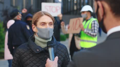 rear view of a caucasian journalist or correspondent in a interview with a caucasian woman who is wearing protective mask in a protest against covid 19