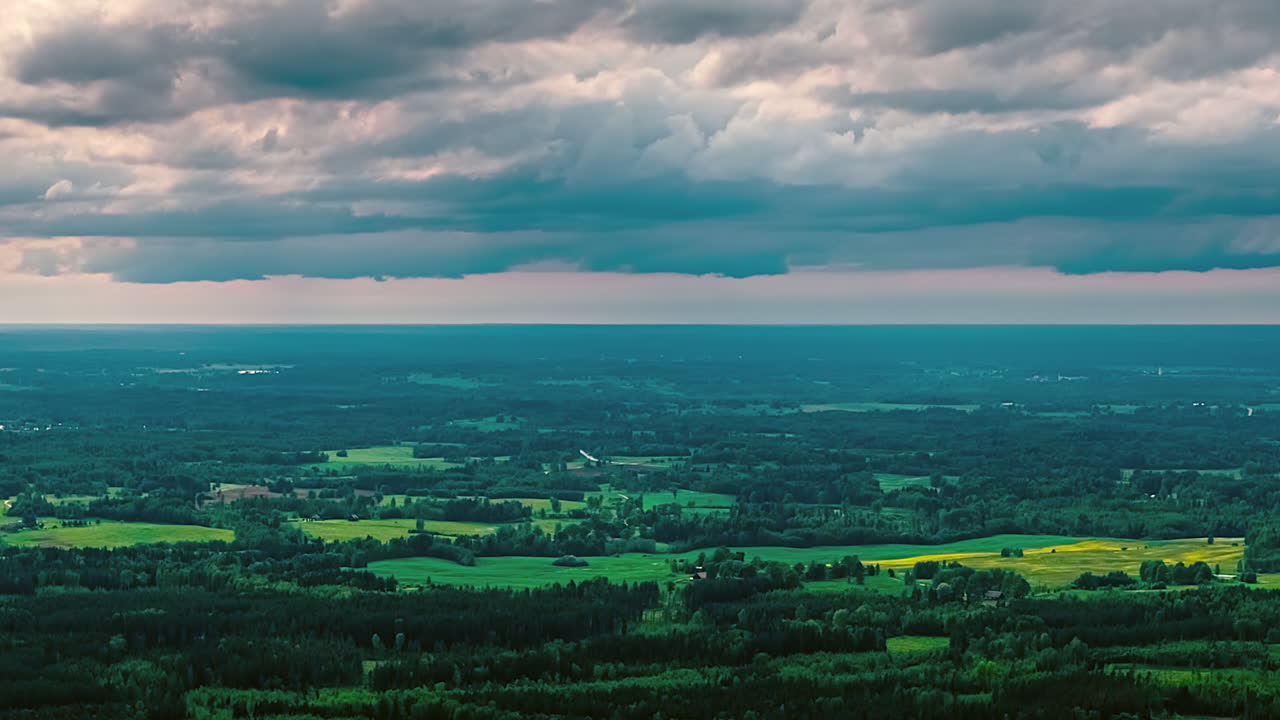 A beautiful time-lapse captures dramatic, moody clouds moving swiftly across the sky at sunset, casting changing light over a vast Latvian landscape of green forests and meadows
