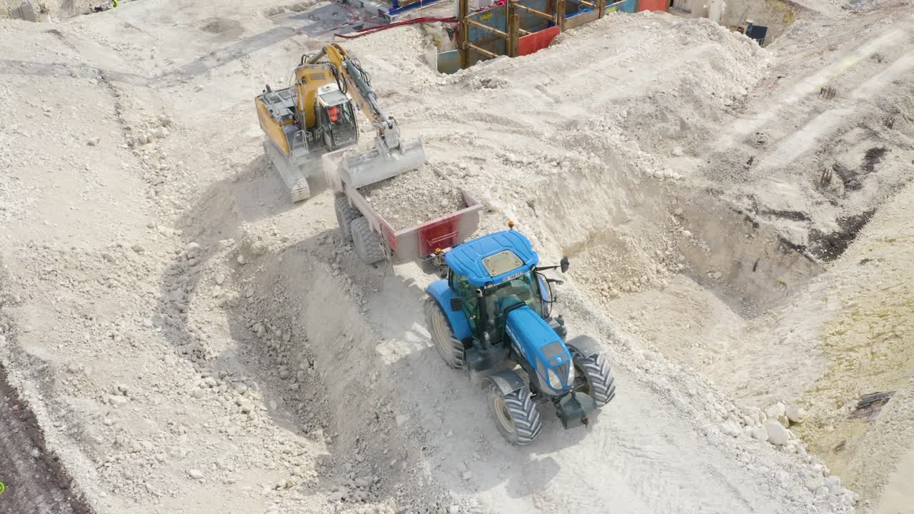 Aerial top down shot over a JCB lifting soil and putting in a tractor at a construction site on a sunny day