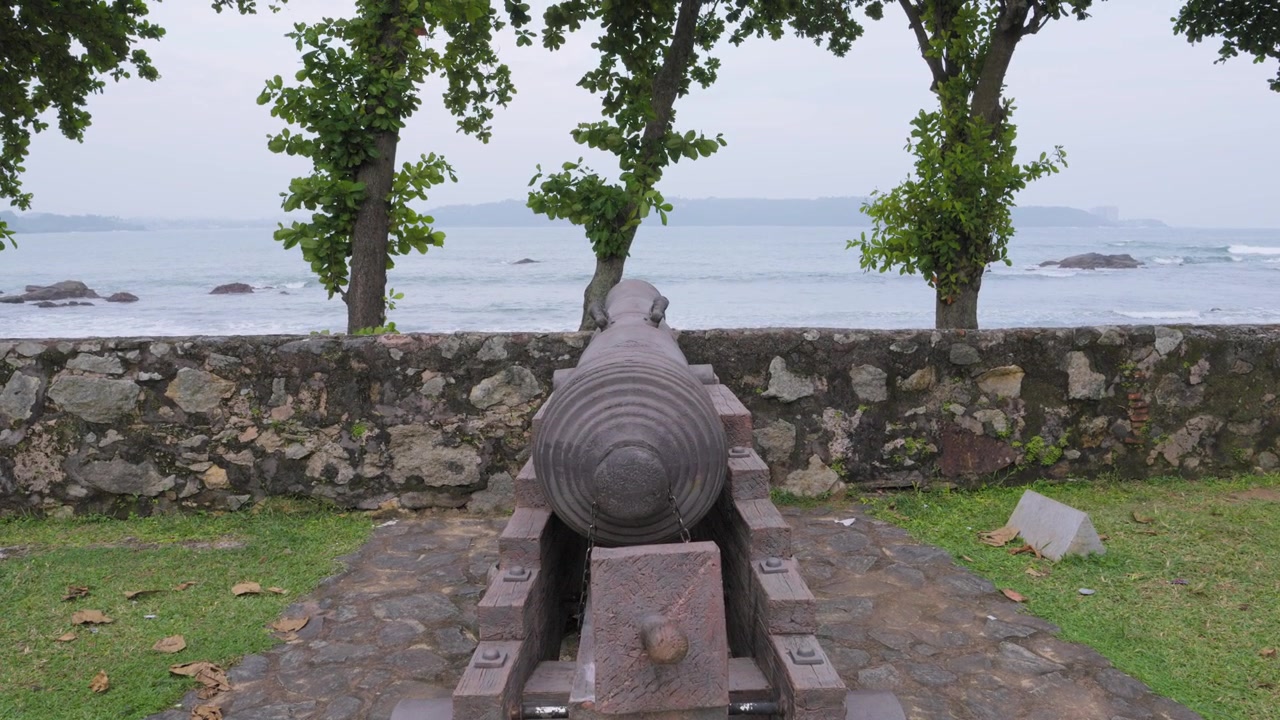 Inquadratura dal basso verso l'alto da un cannone coloniale usurato sulle mura del Forte di Galle, verso alberi, un muro di pietra e onde sfocate dell'Oceano Indiano sulla costa dello Sri Lanka.