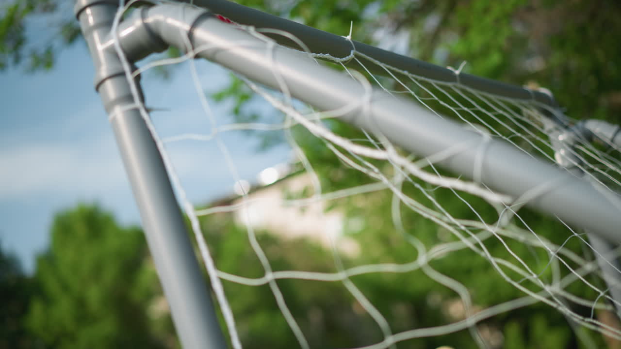 A close-up of a goal post with a ball is shot into the net with precision as the goalkeeper misses, with a blur background of trees