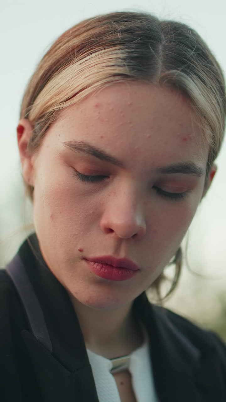 Young lady with neatly parked hair to back appears deeply focused with soft expression while looking downward outdoors with blur background of green trees under clear bright sky