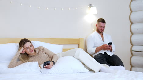 beautiful relaxed woman lies on the double bed looking at her phone. man sits beside on the chair using his gadget.