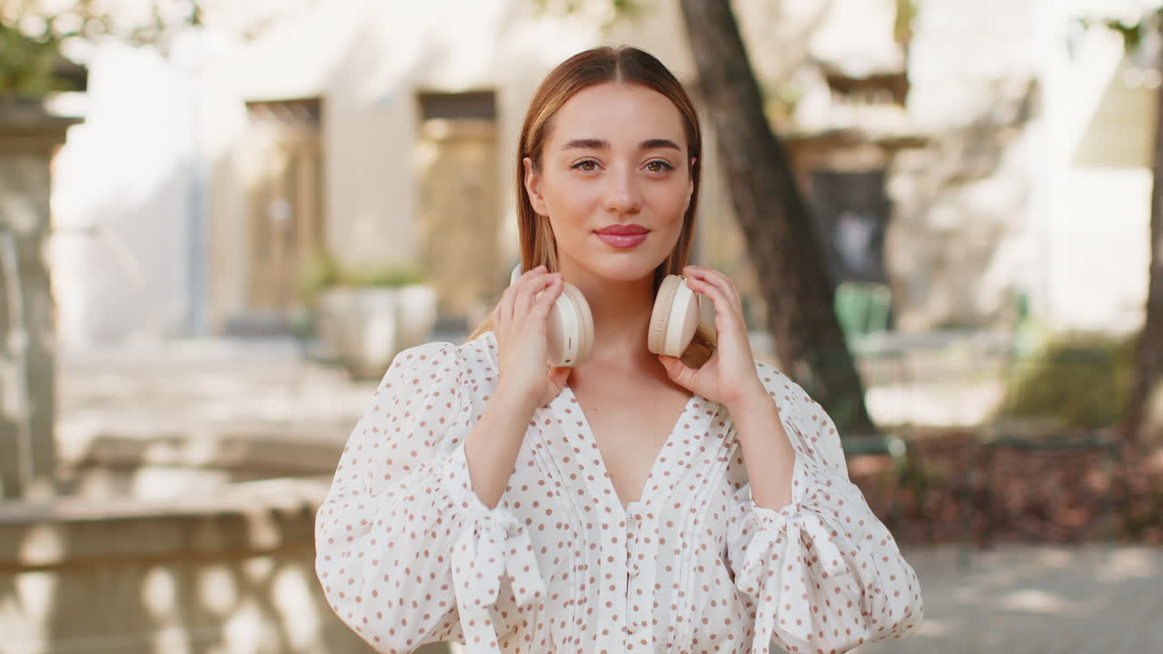 Happy young woman listening music taking off wireless headphones looking at camera smiling outdoors