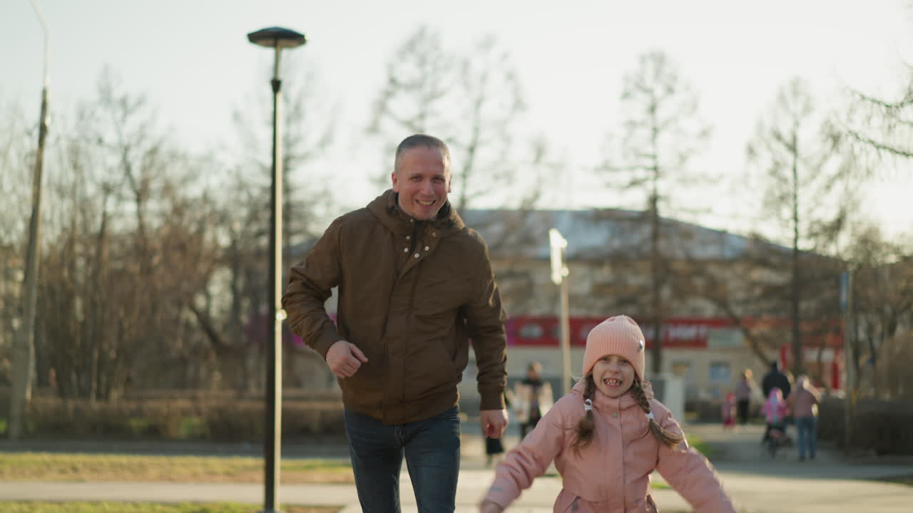 갈색 재과 파란 청바지를 입은 남자가 분홍색 모자와 재을 입은 웃는 어린 소녀와 함께 달리는 즐거운 장면