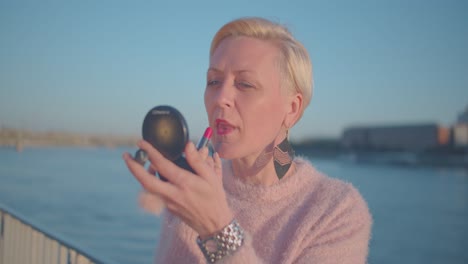 blonde short-haired middle age woman puts red lipstick on lips, close up slomo. healthy lively older woman in outdoors