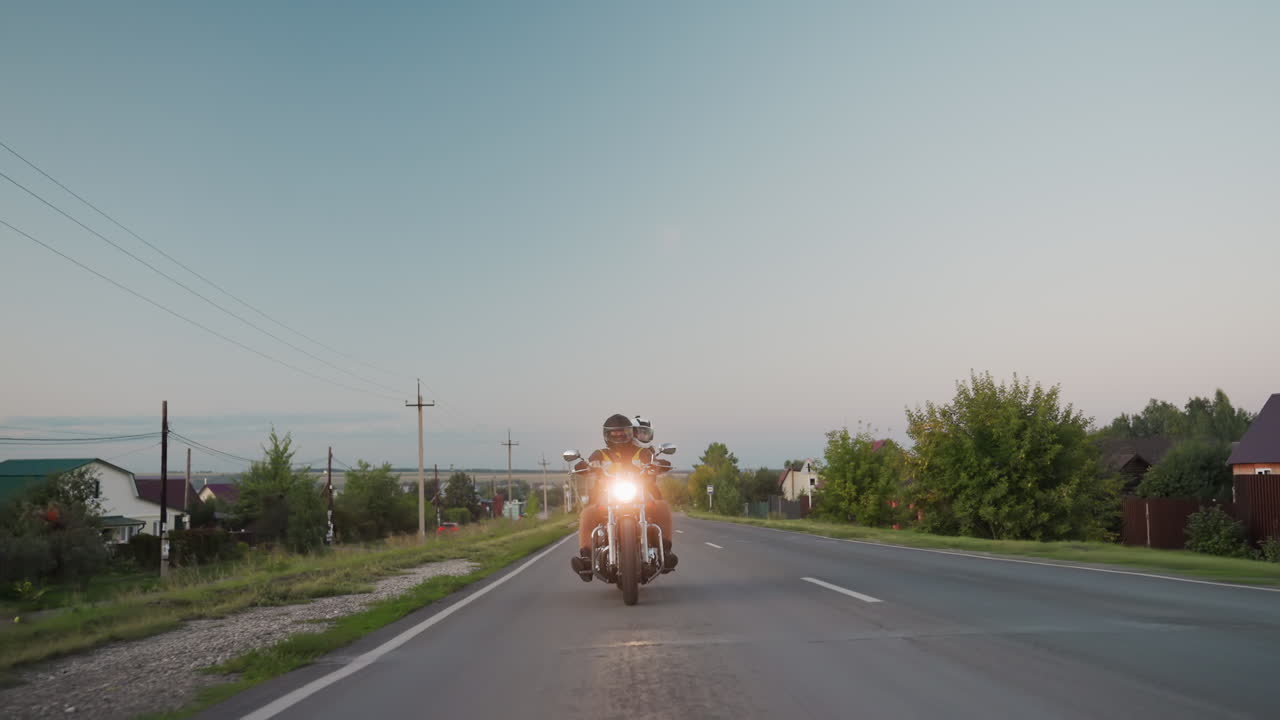 Motorcycle rider with passenger wearing helmets drives countryside road on road trip, passing utility poles, trees, and houses in rural setting under clear evening sky