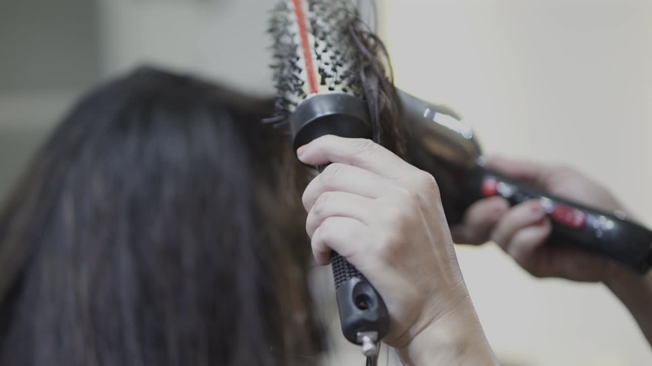 A stylist dries a woman's brown hair using a round brush and hair dryer at a salon.