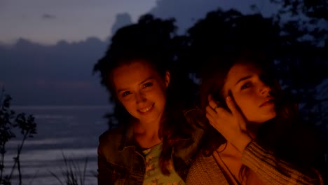 dos jóvenes caucásicas se sientan por la noche junto al fuego en la naturaleza, mirando a la cámara, sonriendo, pensando