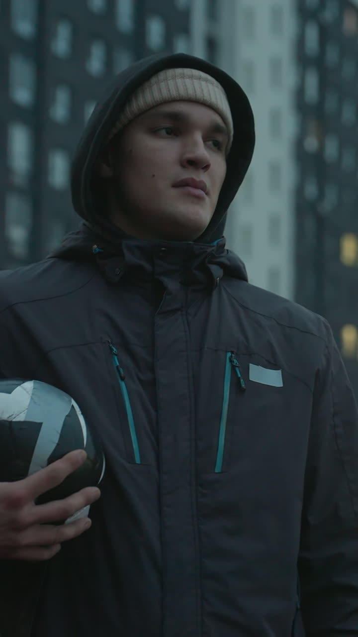 Young man holding soccer ball standing confidently in modern sport arena, surrounded by tall residential buildings under dramatic evening sky, lost in thought