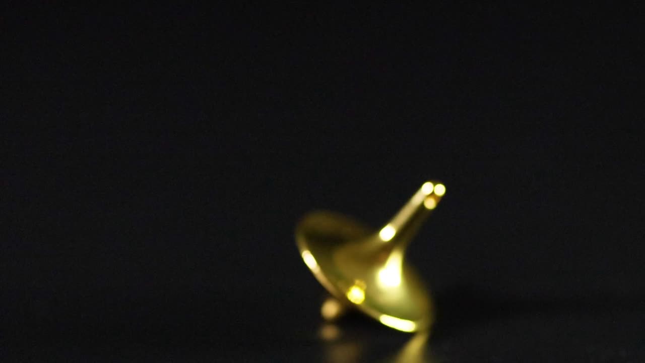 A metallic gold spinning top rotates and wobbles across a dark reflective surface in close-up