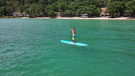 Vista-De-Pájaro-De-Drones-Aéreos-De-ángulo-Bajo-Del-Hombre-Caucásico-Haciendo-Ejercicio-En-Una-Tabla-De-Remo-En-Aguas-Turquesas-Tropicales-Claras,-Con-Playa,-Resort-Y-Costa-En-Tailandia