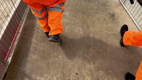 POV-Einer-Gruppe-Von-Füßen,-Die-In-Leuchtend-Orangefarbenen-Warnhosen-Auf-Der-Baustelle-Laufen