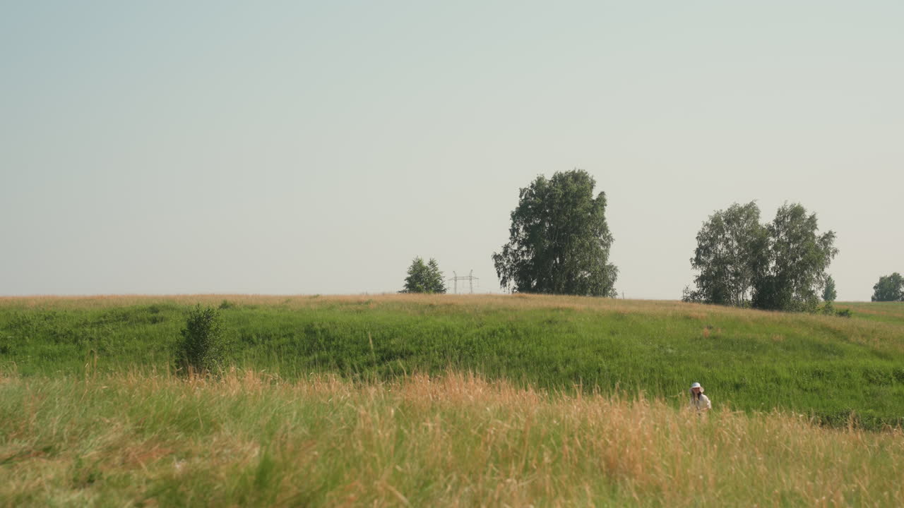 Solitary figure in white hat walks through distant edge of expansive grassy field beneath clear sky, surrounded by layers of wild greenery and clusters of tall trees under calm daylight
