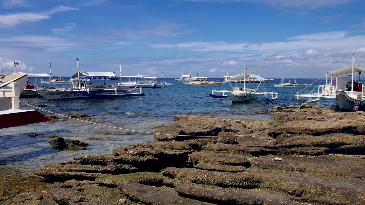 Una sognante scena estiva con diverse imbarcazioni a bilanciere ormeggiate lungo una costa rocciosa sotto un cielo azzurro brillante ad Apo Island, Dauin, Filippine