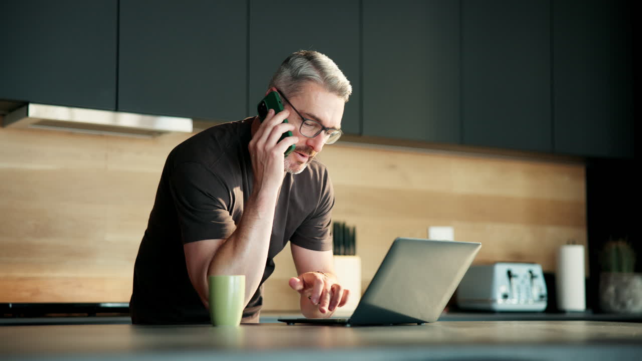 Forretningsmand taler i telefon og arbejder på bærbar computer i et moderne køkken.