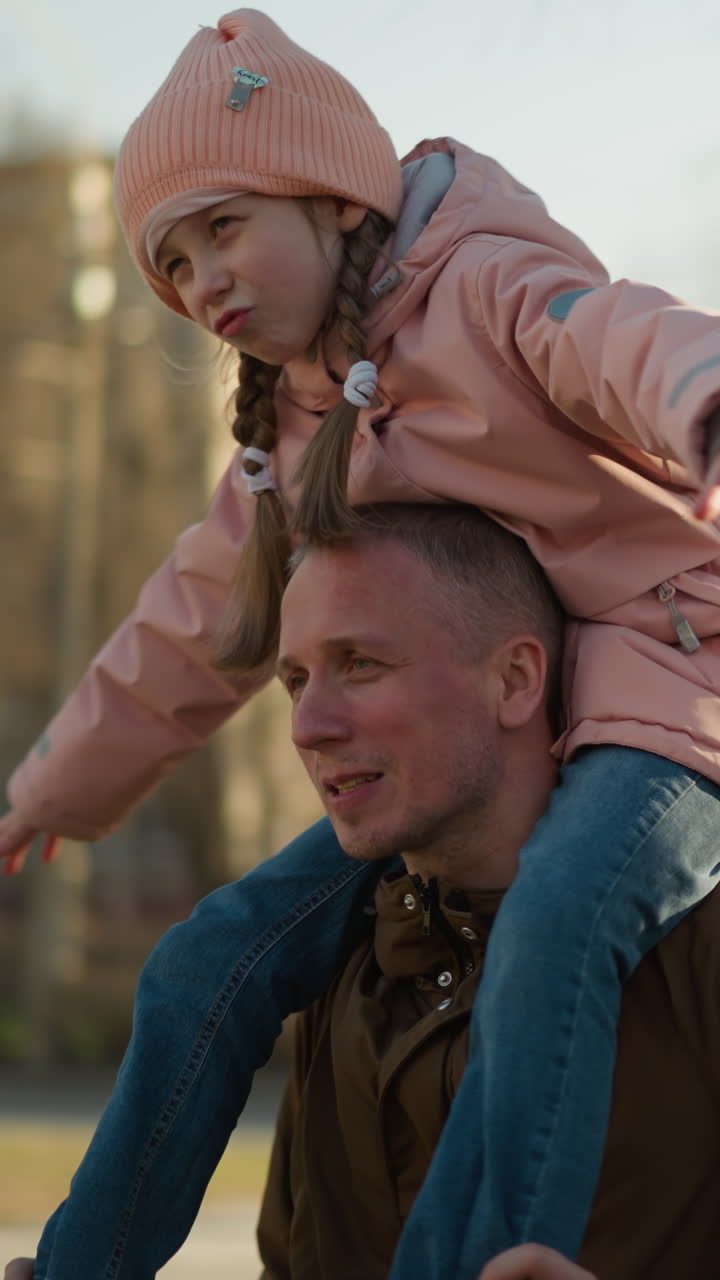 Little girl in a pink cap and jacket joyfully spreads her arms while sitting on her father's neck as the camera circles around them