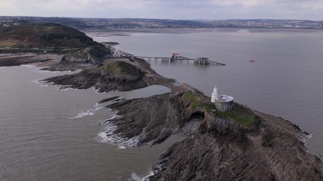 Distant aerial above lighthouse out to Mumbles Pier featuring the Ferris wheel, with ocean waves and a misty coastal atmosphere