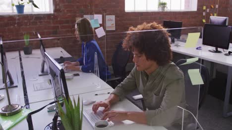 diverse male and female business colleagues sitting at desks with sneeze guard using computers