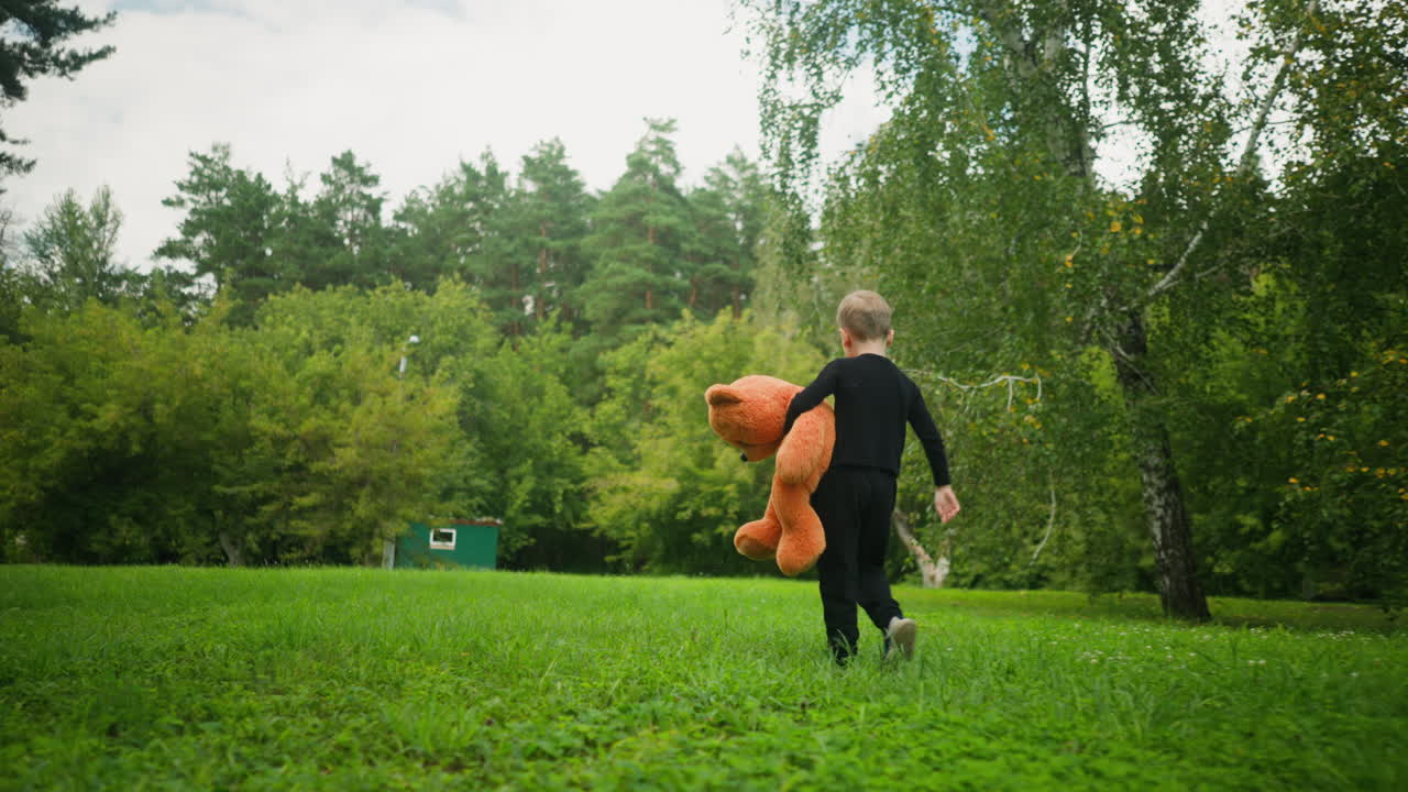 Back view of child in black clothing holding large orange teddy bear while walking playfully across green park surrounded by trees, with small distant building visible in forested background