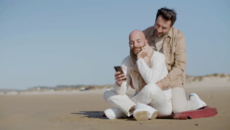 homosexual man sitting on the beach and using mobile phone while his partner approaching and hugging him from back