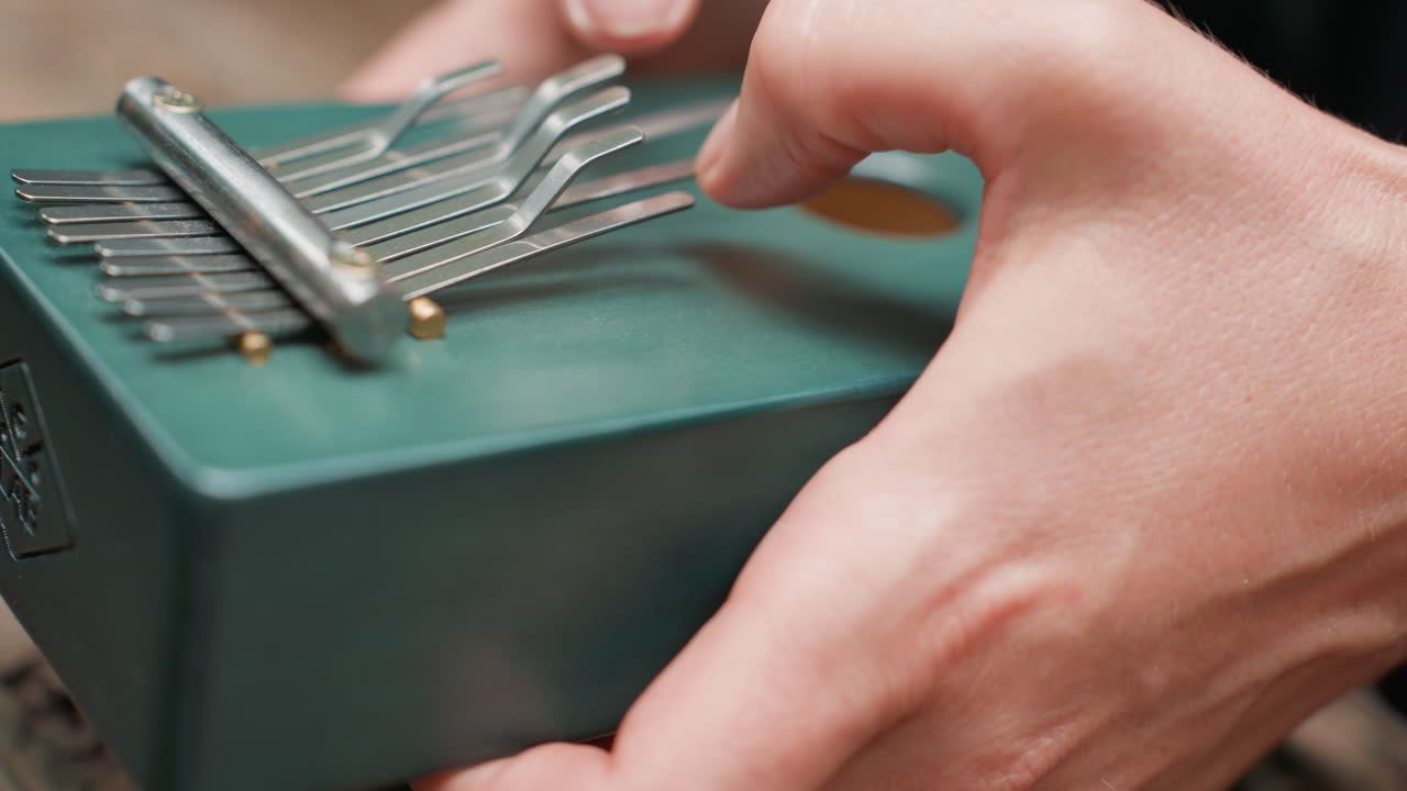 Extreme close up of hands in dark outfit plucking metal tines on small green thumb piano in soft outdoor forest light, focusing on texture, movement, and emotion