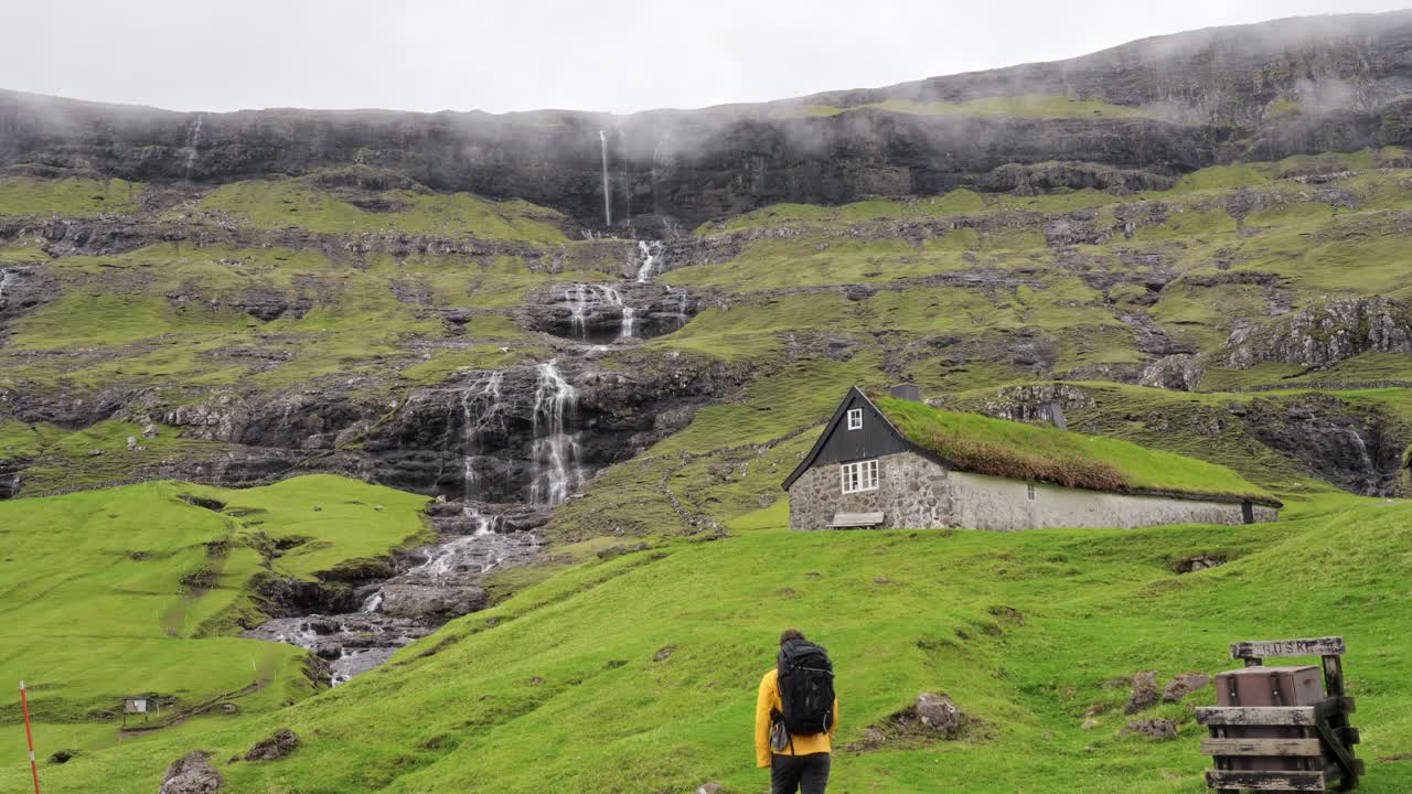 A man walks towards a rustic house near a cliff with cascading waterfalls in Faroe
