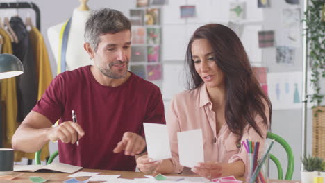 Male-And-Female-Fashion-Designers-At-Desk-In-Studio-Discussing-Colour-Swatches