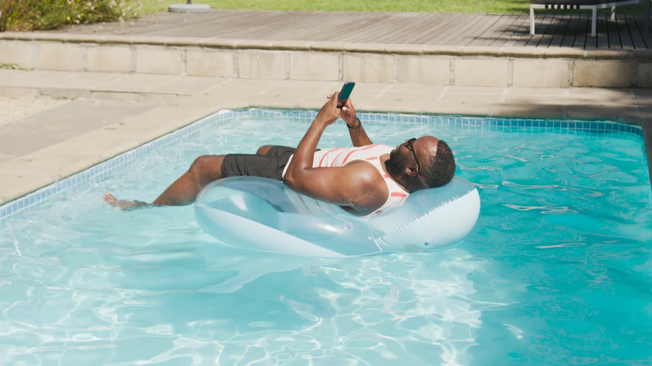 Vertical video: Holding phone, man in white tank nudging blue ring while relaxing in backyard pool