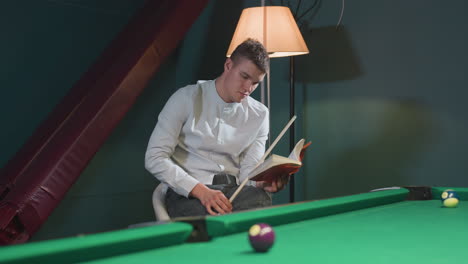 focused young man in white shirt and grey trousers sits on white chair, reading book while holding cue stick near green pool table with scattered billiard balls. soft lighting