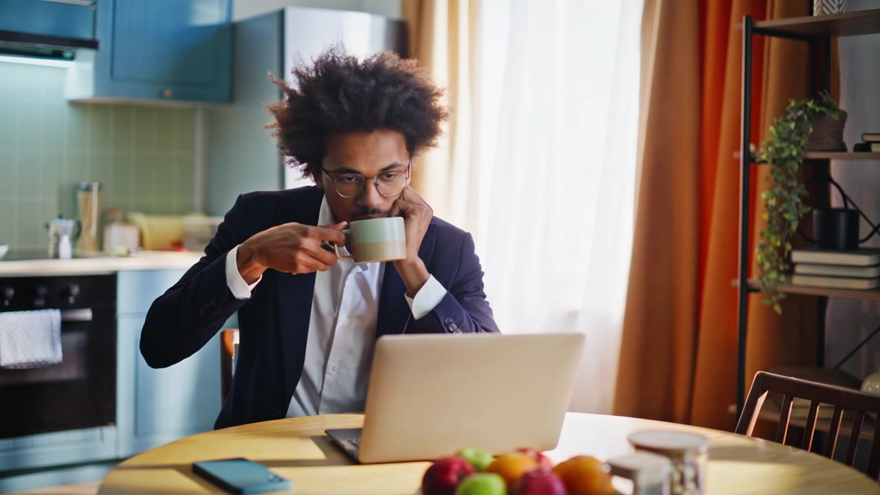 Business man working on laptop in home kitchen, drinking coffee browsing phone