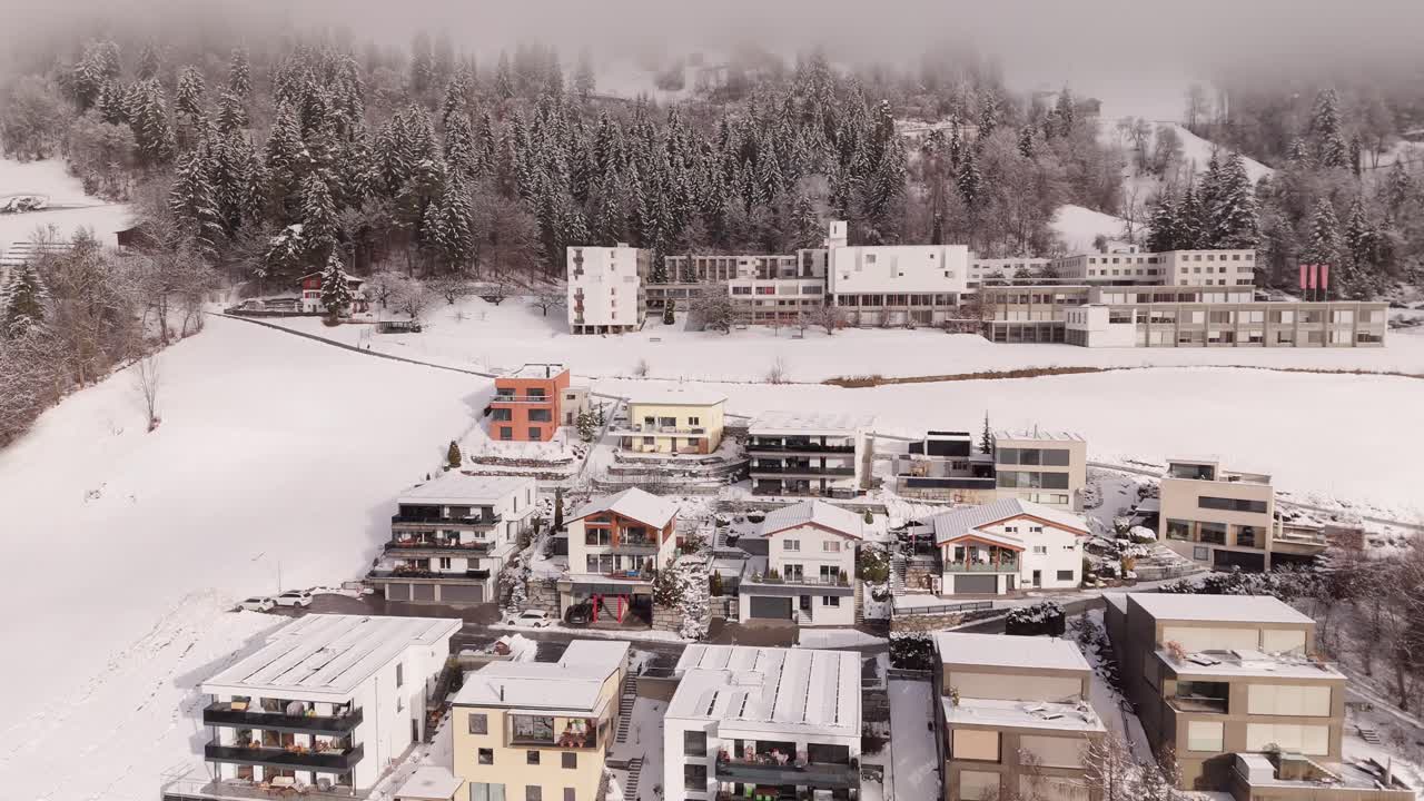 Apartment villas and houses during snowy winter day in Illanz, Switzerland. Aerial Orbit shot. Cloudy day with fog in small swiss town located on snowy hill. Grey cloudscape between conifer trees.