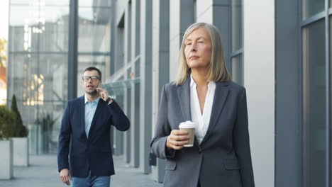 senior woman walking the street with coffee to go in morning, while in the background a businessman is talking on mobile phone